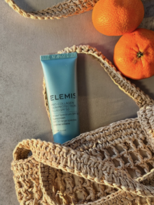 A tube of Elemis Pro-Collagen Skin Protection sunscreen lies on a woven beige bag next to two whole tangerines on a light surface, bathed in warm sunlight. Fearrington Village